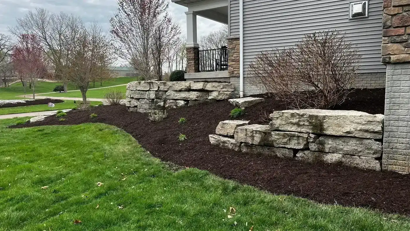 Stacked natural boulder retaining wall with fresh mulch beds beside a house