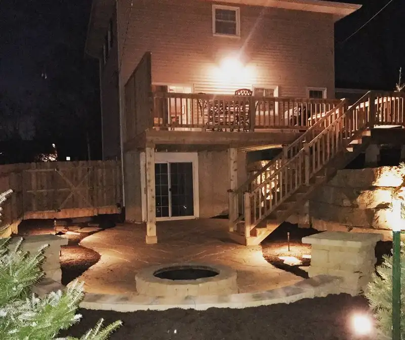 Backyard deck and stone patio lit with landscape lights at night