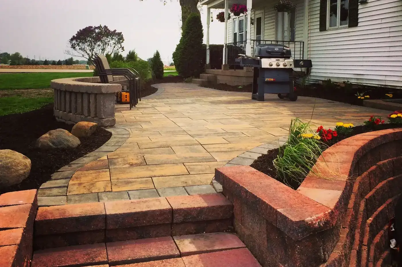 Paver patio with a curved seat wall, pillar caps and planted beds beside a house