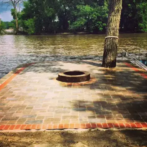 Brick-bordered paver patio with a round fire pit at the edge of a river